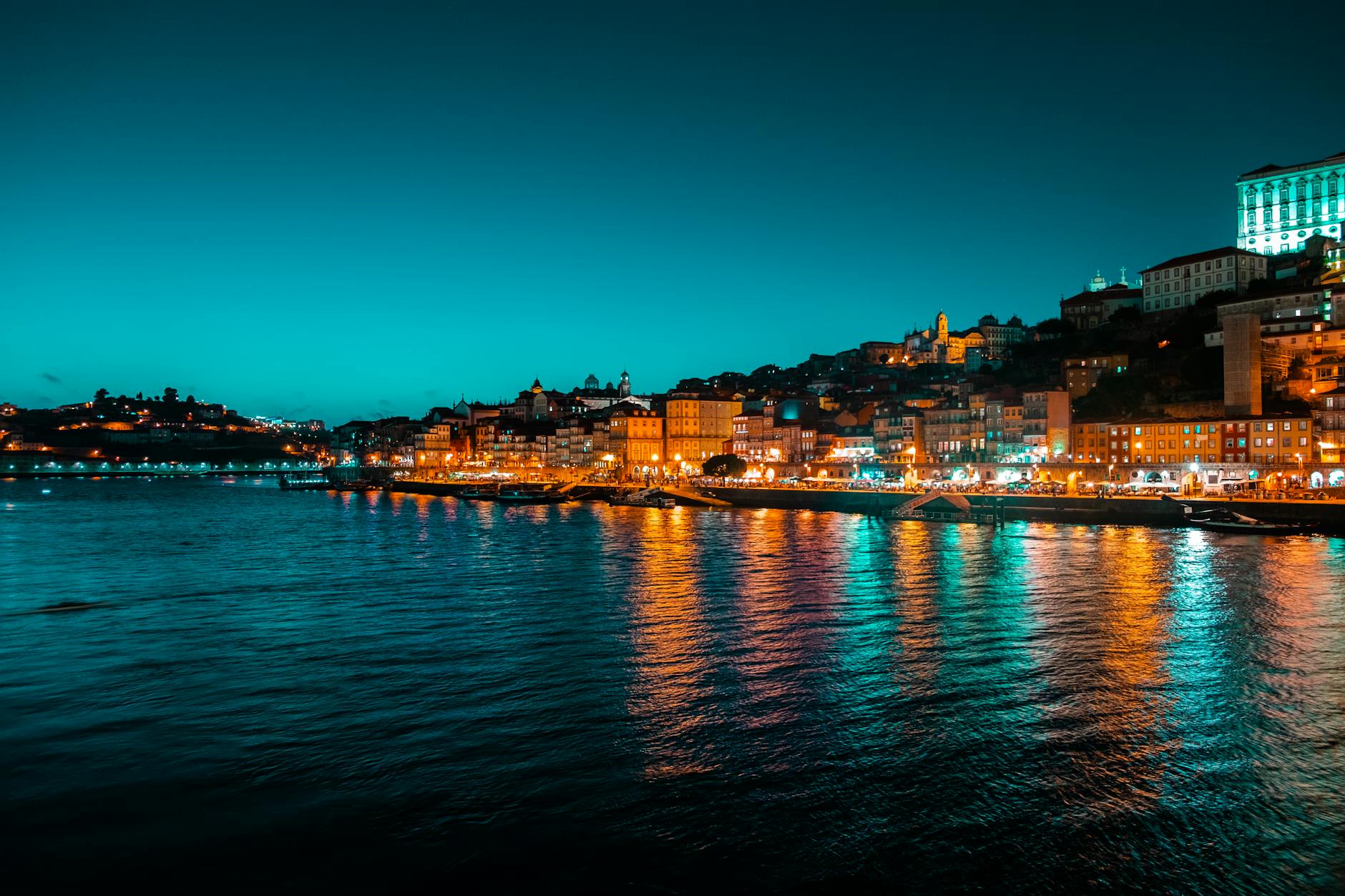 Wieczorna panorama nadrzecznej zabudowy z kolorowymi światłami odbitymi w wodzie Wieczorna panorama nadrzecznej zabudowy z kolorowymi światłami odbitymi w wodzie